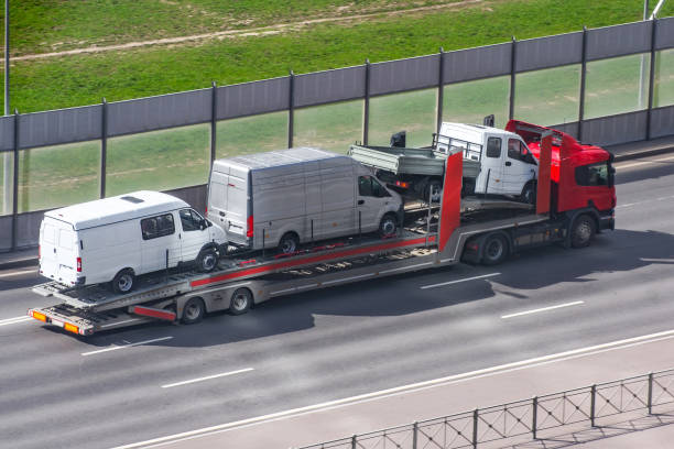A red truck transports three white vans on a flatbed trailer along a divided highway, passing metal sound barriers and green grass, showcasing efficient NC Car Transport Concord services.