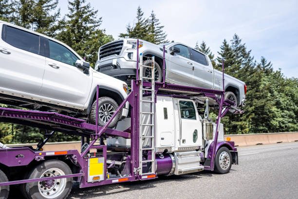 A car carrier truck from NC Auto Transport Concord, NC transports two pickup trucks on a highway, surrounded by tall green trees and a clear sky in the background.