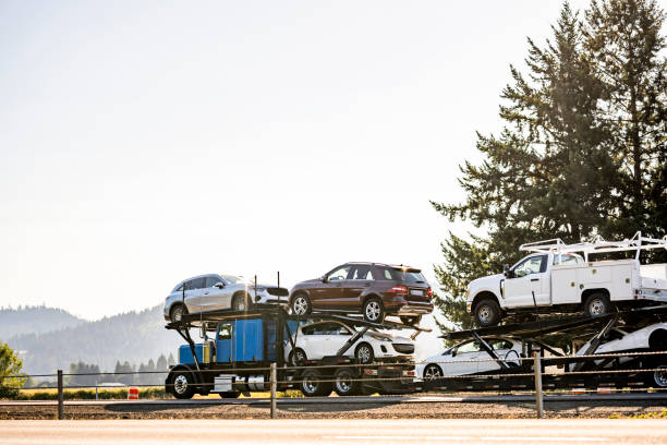 A blue car carrier truck, providing professional NC Car Shipping Concord, transports multiple vehicles, including SUVs and pickup trucks, on a rural road with trees and mountains in the background on a sunny day.