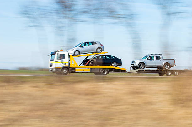 A yellow tow truck hauls three cars—one silver and one black sedan stacked on its bed and a silver pickup on a trailer—on a blurred rural road, suggesting motion and speed, exemplifying the efficiency of Car Transport Concord, NC.