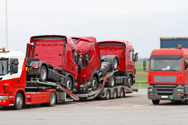 A truck transporter, part of a Car Transport Concord, NC fleet, carries two stacked red truck cabs on a flatbed trailer, parked beside another red truck in an open lot with a cloudy sky.