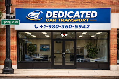 Storefront of Dedicated Car Transport with a blue sign, phone number, and a car logo above glass doors and windows. Conveniently located at the corner of Spring St NW, this trusted provider offers NC Auto Transport Concord services. The office interior is visible through the windows.